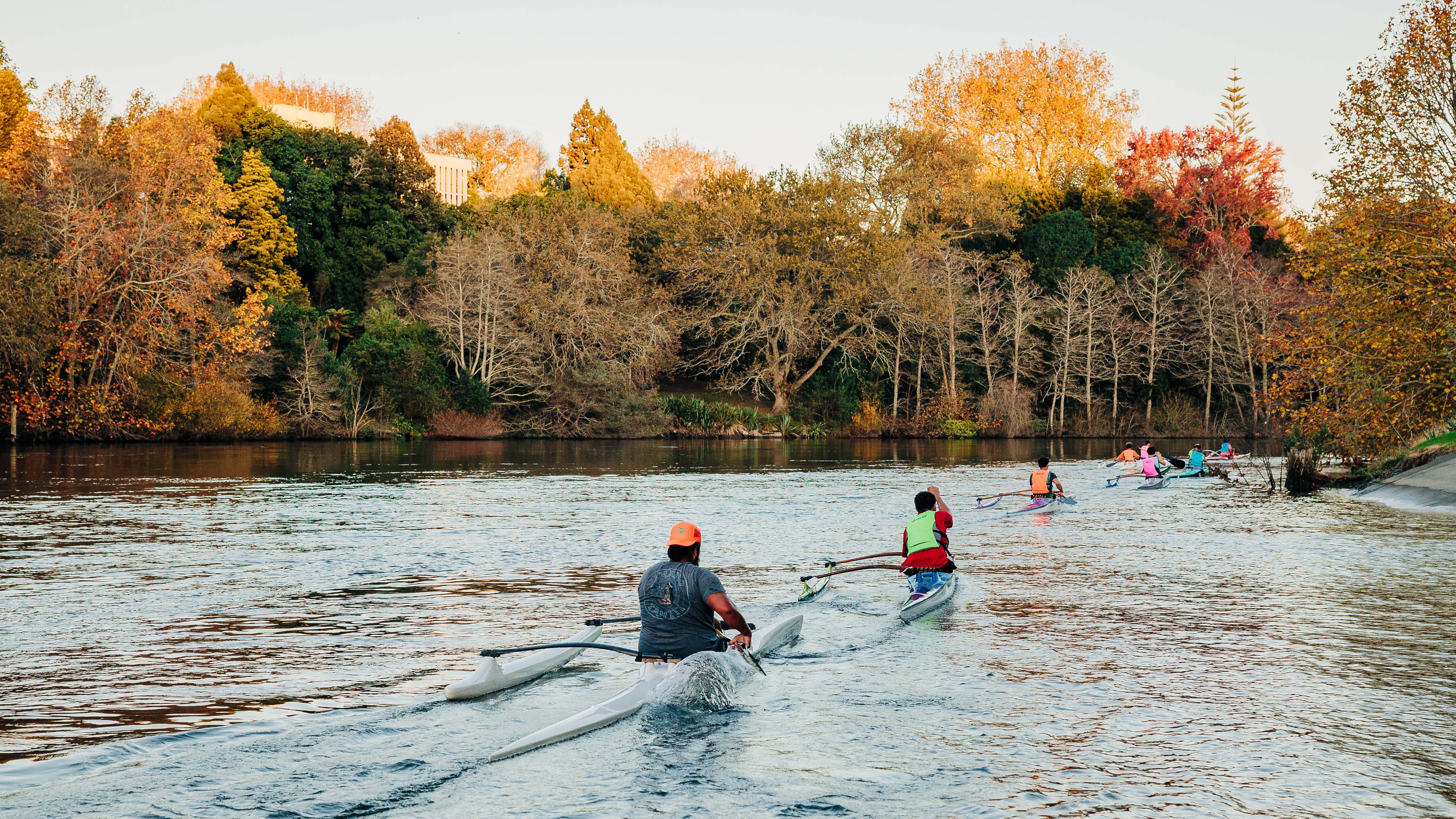 Two people kayaking on a calm stretch of the Waikato River, surrounded by autumn-coloured trees.