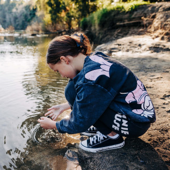 A young person crouching at the edge of a river, dipping their hands into the water.
