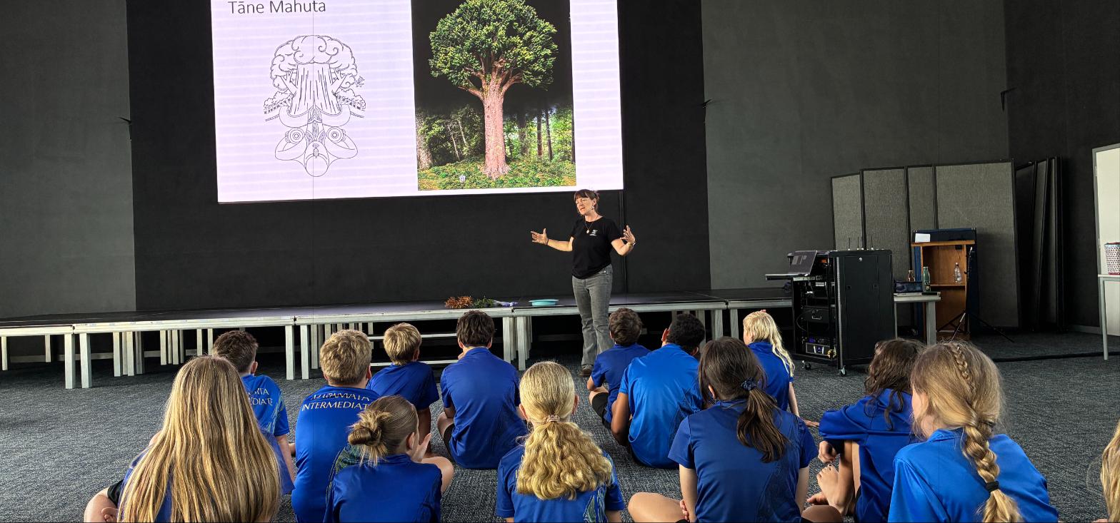 A presenter teaching a group of schoolchildren about kauri trees, with a diagram of a kauri tree and its root system displayed on screen