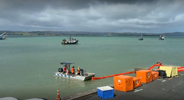 Boat driving away from shoreline pulling an orange 'boom' being fed out of a box on the roadside.